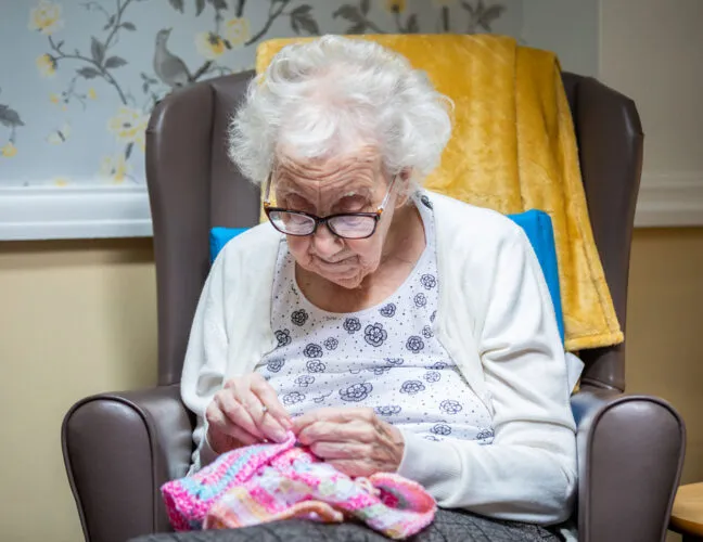 An elderly resident knitting in a comfy chair at care home - Applewood Healthcare